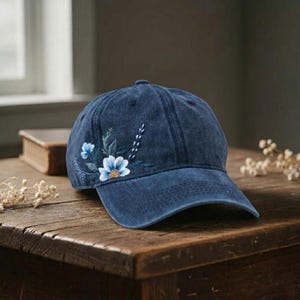 May include: A navy blue baseball cap with a curved brim. The cap features an embroidered floral design in shades of blue, white, and green on the side. The cap is made of a soft, textured fabric.