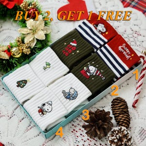 以下が含まれることがあります： クリスマスをテーマにした靴下のギフトボックス。 靴下は白、オリーブグリーン、赤で、スヌーピー、クリスマスツリー、休日のフレーズなど、さまざまな刺繍デザインが施されています。 ボックスはお祝いの要素で飾られています。