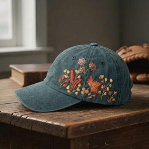 May include: A teal denim baseball cap with embroidered floral patterns in orange, red, and gold. The cap has a curved peak and a button on top. It is displayed on a wooden surface.