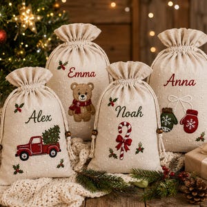 May include: Four beige drawstring gift bags made of fabric, each personalised with embroidered names: Alex, Emma, Noah, and Anna. The bags feature festive designs including a red truck, a teddy bear, a candy cane, and winter mittens.