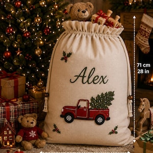 May include: A beige fabric Christmas gift sack featuring the embroidered name Alex, a red truck carrying a green tree, and holly accents. The sack measures 28 inches in height and 21 inches in width, with a drawstring closure and a teddy bear inside.