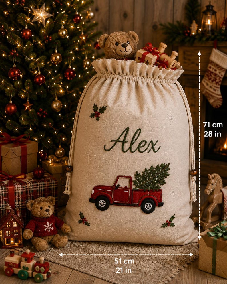 May include: A beige fabric Christmas gift sack featuring the embroidered name Alex, a red truck carrying a green tree, and holly accents. The sack measures 28 inches in height and 21 inches in width, with a drawstring closure and a teddy bear inside.