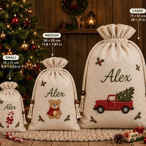 May include: Three beige drawstring gift bags in small, medium, and large sizes. The small bag features a candy cane, the medium a teddy bear, and the large a red truck carrying a tree. Each bag is embroidered with the name Alex and holly.