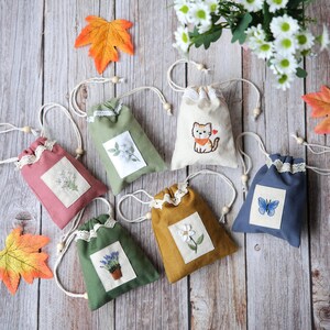以下が含まれることがあります： 刺繍された花柄や動物柄の、小さなカラフルな巾着袋のコレクション。ピンク、グリーン、ブルーなど、さまざまな色のバッグがあり、レースのトリムと木製のビーズで装飾されています。背景は木製の表面です。