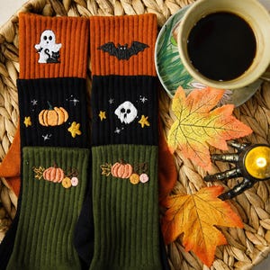May include: A pair of ribbed crew socks featuring colour-blocked sections in orange, black, and olive green. The socks are embroidered with Halloween-themed motifs including a white ghost, a black bat, orange pumpkins, and a white skull.