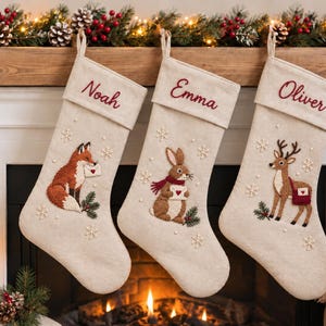 Può includere: Tre calze natalizie beige con animali del bosco ricamati: una volpe, un coniglio e un cervo. Ogni calza è personalizzata con i nomi Noah, Emma e Oliver in corsivo rosso, decorata con fiocchi di neve bianchi e piccole perle.