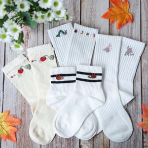 May include: A selection of white and cream socks with embroidered patterns. Designs include strawberries, a caterpillar, flowers, and a sloth. The socks are displayed on a wooden surface, alongside flowers and autumn leaves.