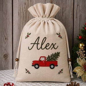 May include: A beige drawstring gift bag featuring embroidered details including the name Alex in green script, a red pickup truck carrying a green pine tree, and small sprigs of holly with red berries in the four corners of the fabric.