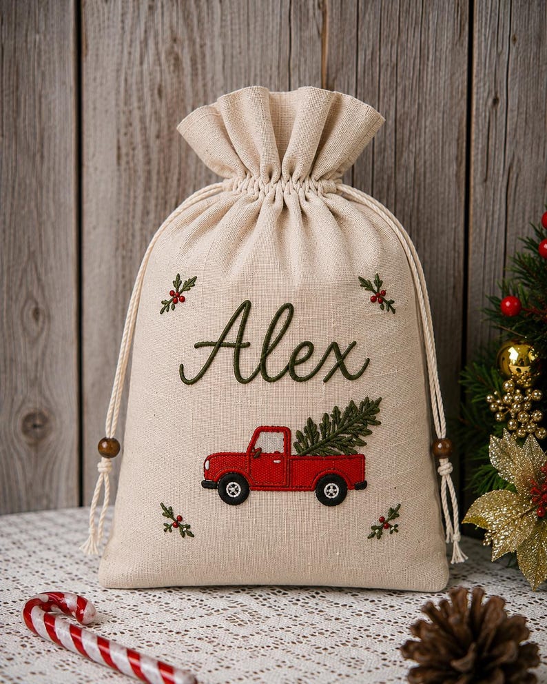 May include: A beige drawstring gift bag featuring embroidered details including the name Alex in green script, a red pickup truck carrying a green pine tree, and small sprigs of holly with red berries in the four corners of the fabric.