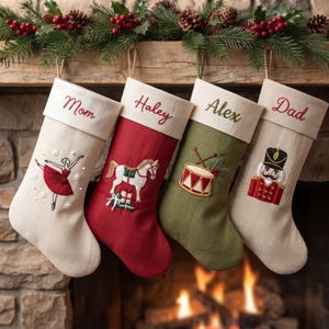 Puede incluir: Cuatro calcetines navideños personalizados colgados en fila. El primero es crema con una bailarina roja bordada, el segundo es rojo con un caballo blanco y regalos, el tercero es verde oliva con un tambor y el cuarto es crema con un cascanueces.