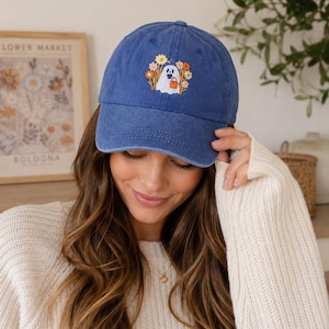 May include: A blue baseball cap featuring an embroidered design of a white ghost holding a small orange pumpkin bucket, surrounded by a colourful arrangement of yellow, orange, and white flowers and tall grasses on the front panel.