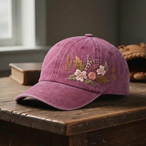 May include: A dusty rose baseball cap with embroidered floral design. The cap features a curved brim and a fabric strap closure. The floral embroidery includes light pink, peach, and brown flowers with green and brown leaves.