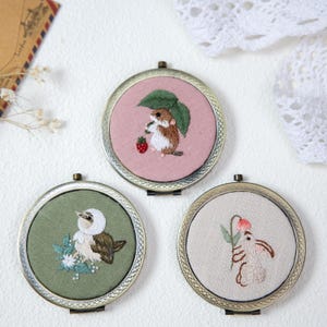 Könnte beinhalten: Drei runde Kompaktspiegel im antiken Stil. Jeder Spiegel hat ein gesticktes Design: ein Eichhörnchen mit einer Erdbeere, ein Vogel mit Blumen und ein Kaninchen mit einer Blume. Die Spiegel haben einen bronzefarbenen Metallrahmen.