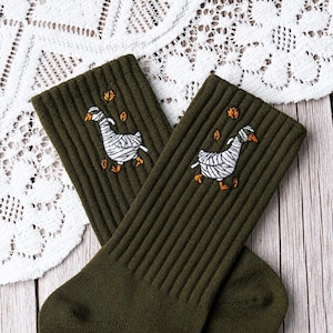Könnte beinhalten: Ein Paar olivgrüne Rippstricksocken mit einem gestickten Motiv einer weißen Gans, die wie eine Mumie in Bandagen gewickelt ist, akzentuiert durch kleine orangefarbene Herbstblätter, die um die Gans auf jeder Socke verstreut sind.