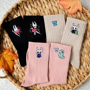 Puede incluir: Una colección de calcetines coloridos con diseños bordados. Los calcetines son negros, beige y rosas, con varios personajes de dibujos animados y diseños de mariposas. Los calcetines están colocados en una cesta de mimbre con hojas de otoño.