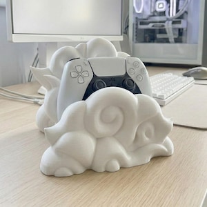 Pode incluir: Um suporte branco em forma de nuvem para comando de jogos com um comando PlayStation 5 dentro. O suporte está numa secretária de cor clara, com um monitor de computador, um teclado e um rato no fundo. O comando é branco e preto.