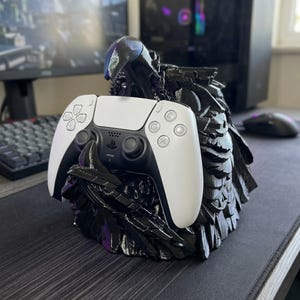 Puede incluir: Un mando de PlayStation 5 en blanco y negro descansa en un soporte negro texturizado. El soporte tiene un diseño esculpido único. Un monitor de ordenador, un teclado y un ratón son visibles en el fondo.