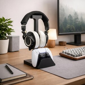 May include: A black headphone stand with a white and black gaming controller base. The stand holds a pair of black and white headphones. The desk setup includes a computer monitor, keyboard, and a small plant in a pot.