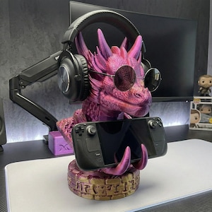 May include: A pink and purple dragon-shaped gaming accessory stand. The dragon wears sunglasses and holds a black handheld gaming device. Headphones rest on the dragon's head. The stand has a decorative base.