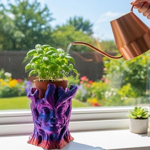 May include: A basil plant in a brown pot is placed in a decorative planter. The planter is a face-shaped design with purple, blue, and red hues. A copper watering can is pouring water onto the plant. The scene is set on a windowsill with a garden view.