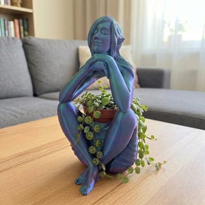 Puede incluir: Una maceta con forma de figura sentada en azul y morado iridiscente sostiene una pequeña maceta marrón con una planta suculenta colgante. La figura tiene los ojos cerrados y orejas puntiagudas. La maceta está sobre una mesa de madera clara.
