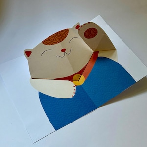 May include: A folded paper card depicting a smiling, beige cat with a raised paw. The cat has brown patterned markings and a red collar with a gold charm. The cat is wearing a blue garment, set against a white background.