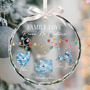 May include: A round clear glass ornament with a white ribbon, featuring the text "FAMILY LOVE Knows No Distance". It displays three stained-glass style state maps of Ohio, Washington, and New York, each with a red heart, hanging from colorful Christmas lights.