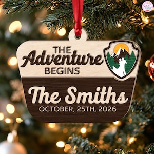 May include: A wooden ornament with a red ribbon featuring the text THE ADVENTURE BEGINS, The Smiths, and OCTOBER, 25TH, 2026. It includes a shield-shaped graphic showing a silhouette of a couple against a mountain landscape with a yellow sun.