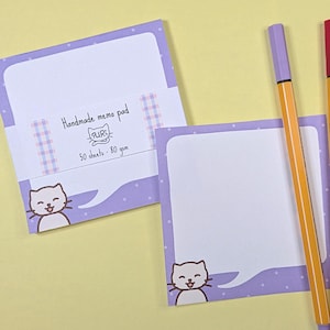 May include: A handmade memo pad with a cat illustration, 50 sheets, 80 gsm, and a purple polka dot design. Two orange and purple pens and a red and orange pen are next to the memo pad on a yellow background.