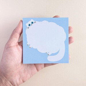 Puede incluir: Una nota adhesiva cuadrada de color azul claro con la ilustración de un gato blanco grande y esponjoso. El gato tiene un cuerpo redondo, una cola larga y una cara pequeña con ojos de color azul brillante y una boca diminuta.