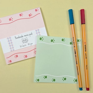 May include: A pink memo pad with paw print designs and the text "Handmade memo pad". A light green memo pad with paw print designs and two orange pens with blue and red tips are also present.