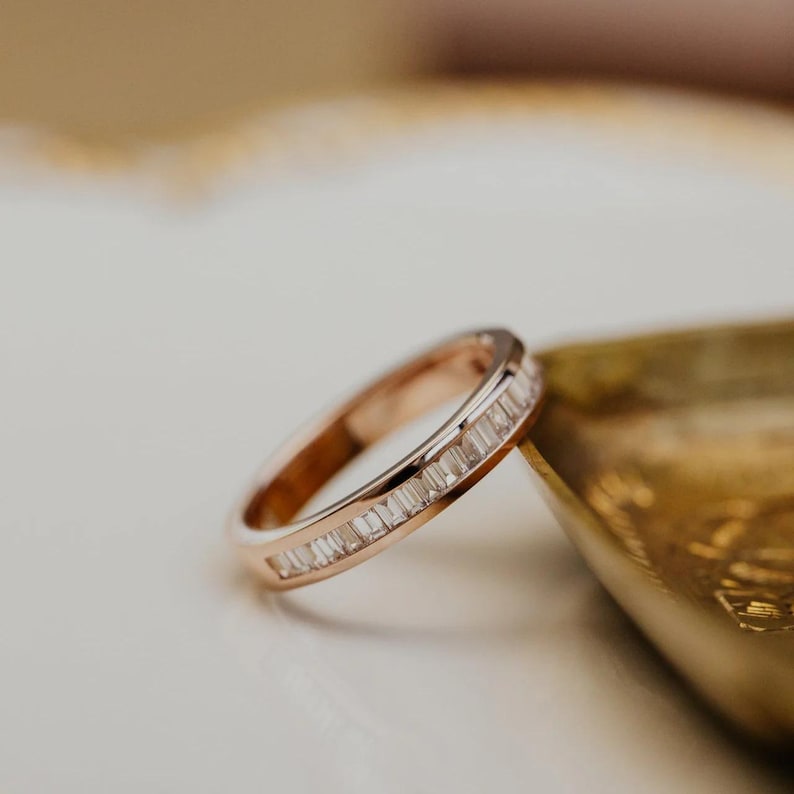 May include: Rose gold band ring with a row of rectangular-cut, clear gemstones. The ring is set on a white surface next to a gold-colored dish. The ring's design is simple and elegant.