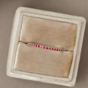 May include: A delicate gold ring set with small, vibrant red gemstones. The ring is displayed in a beige velvet jewelry box, showcasing its elegant design and craftsmanship. The gemstones are evenly spaced, creating a continuous line of color.