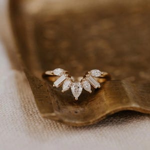 Op de afbeelding: Een gouden ring met een gebogen ontwerp, met een centrale peervormige diamant en verschillende baguette- en marquise-geslepen diamanten. De ring ligt op een goudkleurig oppervlak, dat de ingewikkelde details en schittering laat zien.
