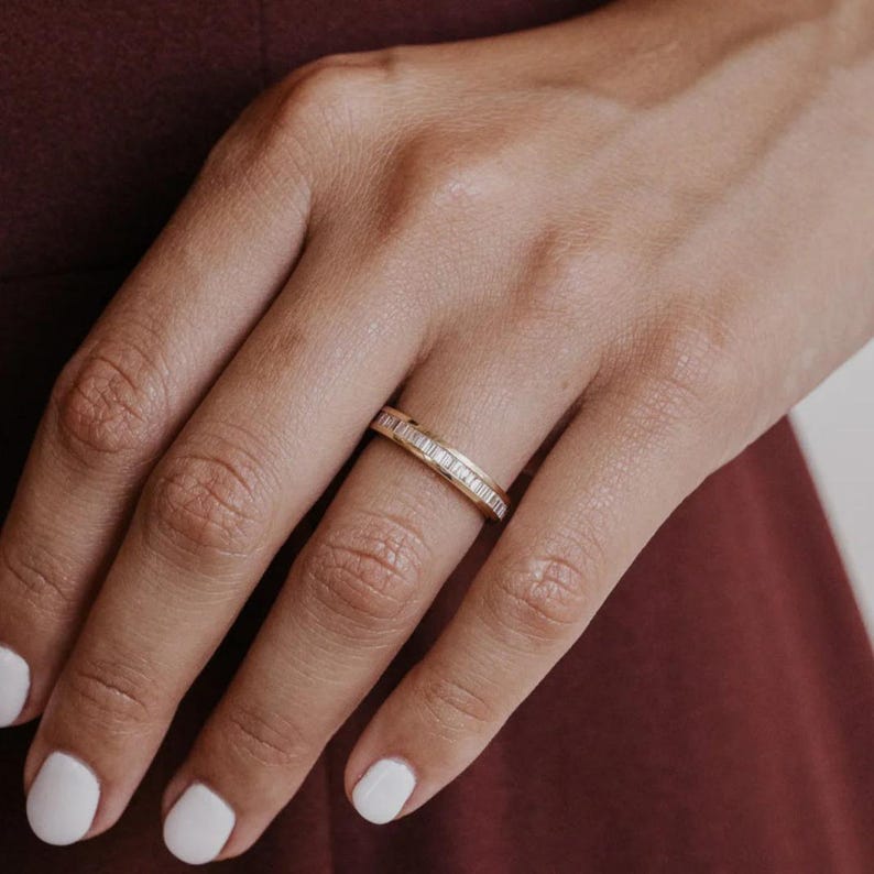 May include: A gold band ring with a row of small, rectangular-cut diamonds. The ring is on a finger, with a burgundy background. The ring's design is simple and elegant, suitable for everyday wear or special occasions.