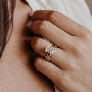 May include: A close-up of a hand wearing two gold rings. The primary ring features a large, emerald-cut diamond. A second ring, also gold, is set with smaller diamonds. The rings are set against a soft, neutral background.