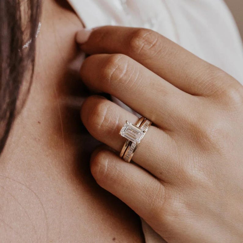 May include: A close-up of a hand wearing two gold rings. The primary ring features a large, emerald-cut diamond. A second ring, also gold, is set with smaller diamonds. The rings are set against a soft, neutral background.