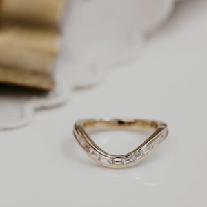 Puede incluir: Un anillo de oro curvado con una fila de piedras preciosas rectangulares y transparentes. El anillo tiene un diseño en forma de V y está colocado sobre un fondo blanco. Probablemente sea una alianza de boda o un anillo de moda.