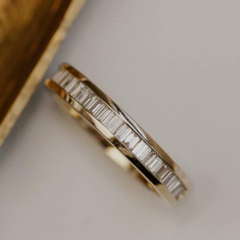 May include: A gold band ring with a row of rectangular-cut, clear gemstones. The ring is set against a neutral background, with a portion of a gold-colored tray visible in the upper left corner.