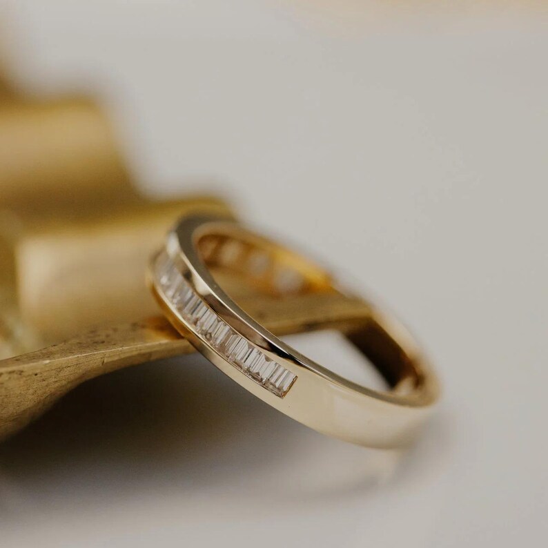 May include: A gold band ring with a row of rectangular-cut gemstones. The ring is set on a gold-colored surface, with a white background. The ring is a close-up shot, showcasing the details of the gemstones and the band.
