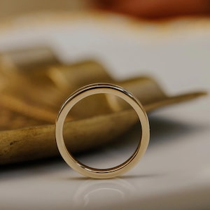 May include: A gold wedding band with a polished finish. The ring is circular and sits on a white surface with a wooden leaf-shaped object in the background. The ring is a symbol of commitment and love.