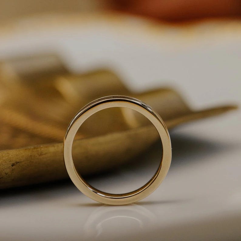 May include: A gold wedding band with a polished finish. The ring is circular and sits on a white surface with a wooden leaf-shaped object in the background. The ring is a symbol of commitment and love.