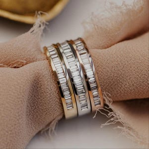 May include: Four rings with baguette-cut gemstones in gold, silver, and rose gold tones. The rings are stacked together and rest on a light brown fabric. The gemstones sparkle, reflecting light. The rings are a jewelry item.