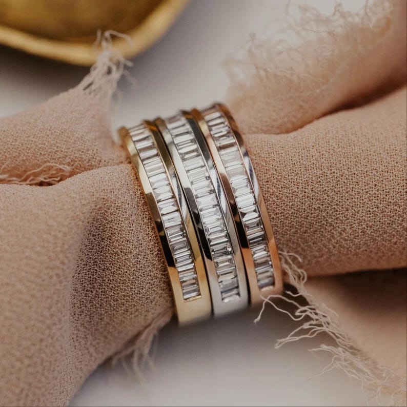 May include: Four rings with baguette-cut gemstones in gold, silver, and rose gold tones. The rings are stacked together and rest on a light brown fabric. The gemstones sparkle, reflecting light. The rings are a jewelry item.