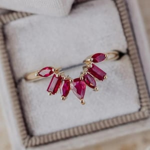 May include: A gold ring featuring a fan-shaped design of vibrant red gemstones. The gemstones are cut in various shapes, including marquise, baguette, and pear, and are set in a delicate, gold band. The ring is displayed in a gray jewelry box.