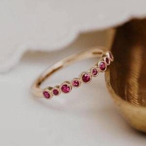 Peut inclure: Une bague en or ornée d'une rangée de sept petites pierres précieuses rondes d'un rose vif, serties individuellement. Chaque pierre est entourée d'une délicate texture milgrain, formant une ligne continue de couleur sur le dessus de la bague.