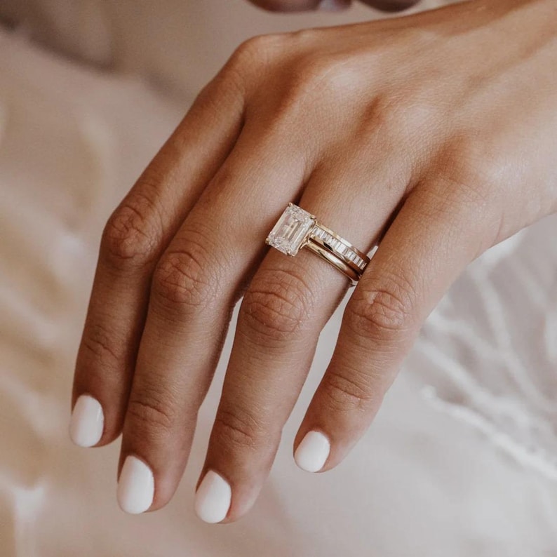 May include: A gold engagement ring with a large, rectangular-cut diamond is displayed on a hand with white nail polish. A second gold band with smaller diamonds sits below it. The rings are set against a soft, blurred background.
