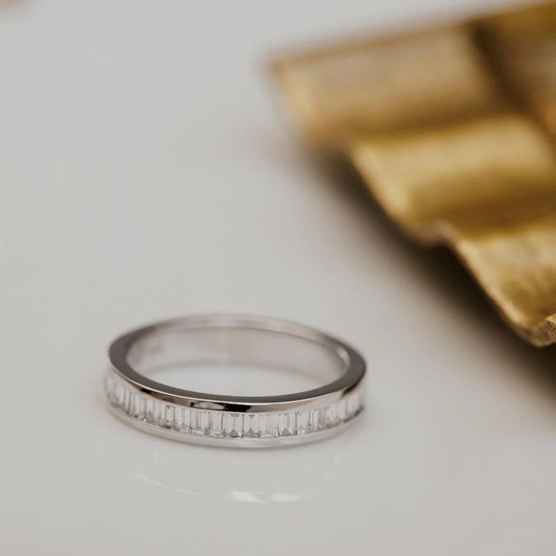 May include: A silver ring with a band of rectangular, clear gemstones. The ring is set on a white surface, with a gold-colored, textured object in the background. The ring's design is simple and elegant.