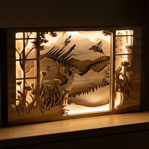 Peut inclure: Diorama en bois éclairé représentant un paysage de montagne avec un cerf. La conception en couches crée de la profondeur, avec des arbres et des fleurs encadrant la scène. La lumière chaude émane de l'intérieur, créant une ambiance chaleureuse.
