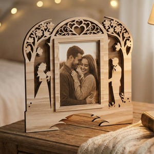 Peut inclure: Cadre photo en bois avec un motif découpé au laser représentant un cœur, des fleurs et des silhouettes d'une mariée et d'un marié. Le cadre contient une photographie sépia d'un couple. Le cadre est sur une surface en bois.